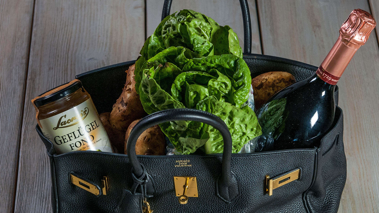 Das Bild zeigt eine luxuriöse schwarze Handtasche, die mit hochwertigen Lebensmitteln gefüllt ist: ein Glas Geflügelfond der Marke Lacroix, frischer grüner Salat, Kartoffeln und eine Flasche Champagner mit roséfarbener Folie. Das Foto symbolisiert Genuss, Qualität und exklusive Feinkostprodukte.